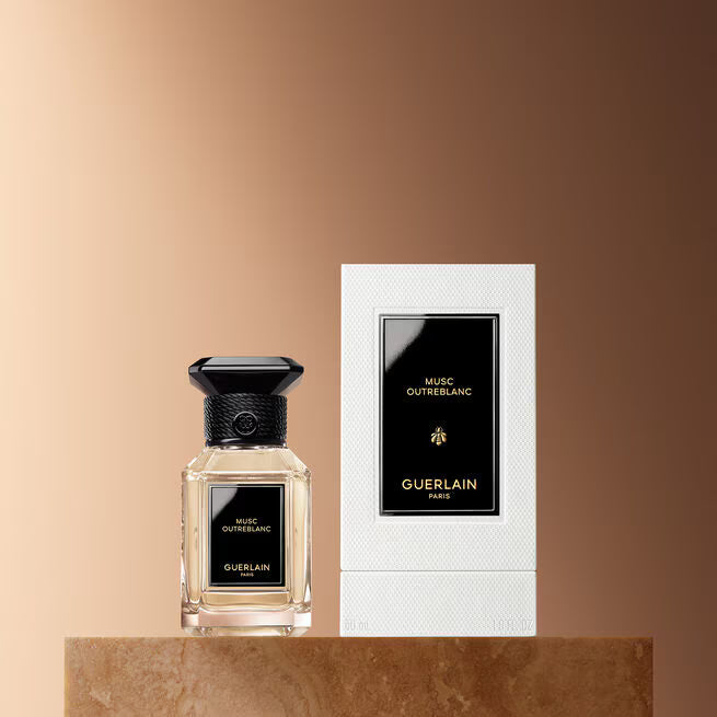 A bottle of Guerlain Music Outreblanc perfume with a black cap stands next to its white textured packaging box, set on a brown stone pedestal against a warm beige gradient background.